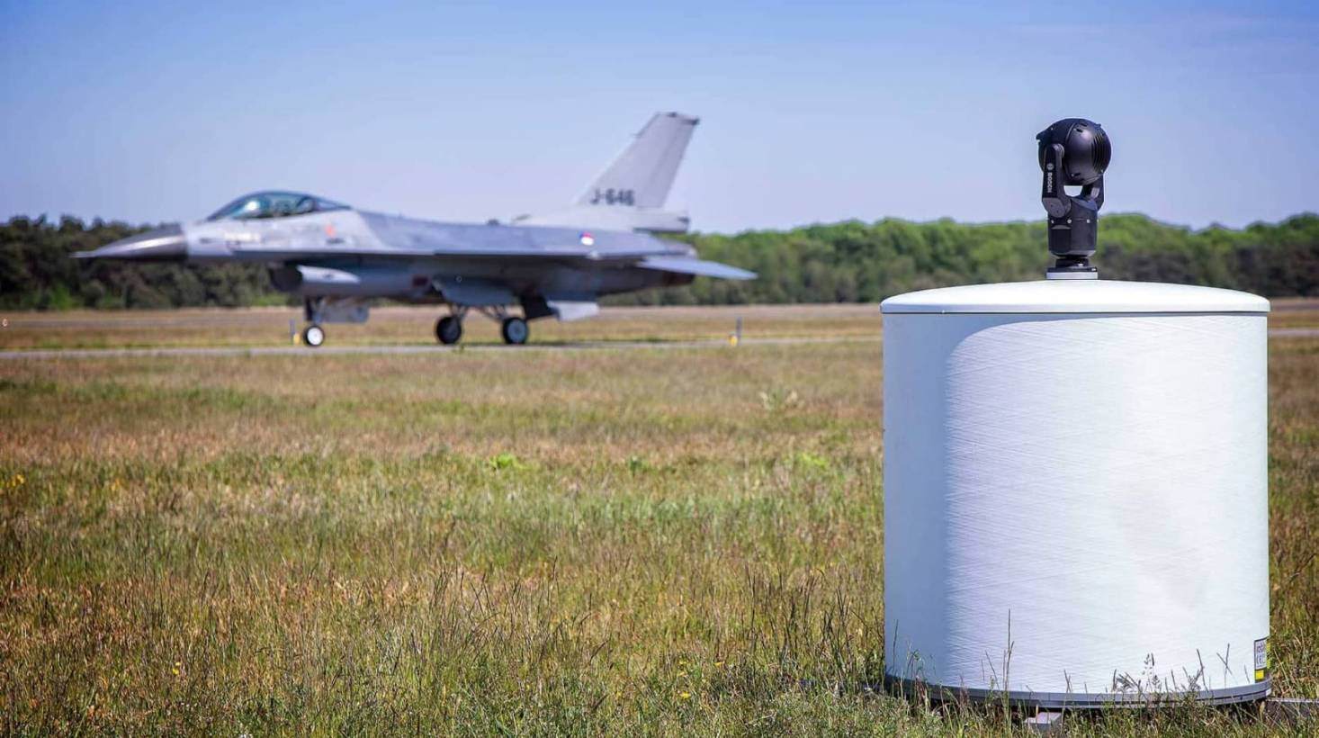 ELVIRA drone detection radar mounted on a white cylinder beside a runway, with a gray fighter jet parked in the background