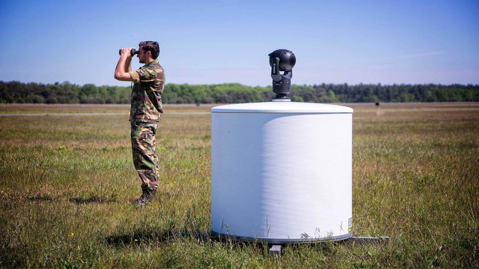 Soldier in green camo standing in a field, using a pair of binoculars to track a drone spotted by the ELVIRA radar (mounted on a white platform) behind him